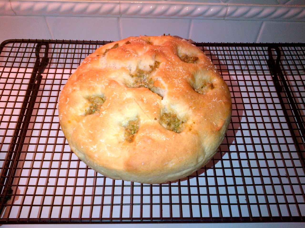 A golden-brown round homemade focaccia bread cooling on a black wire rack. The bread has dimples and is topped with a sprinkle of herbs, likely rosemary, for added flavor.