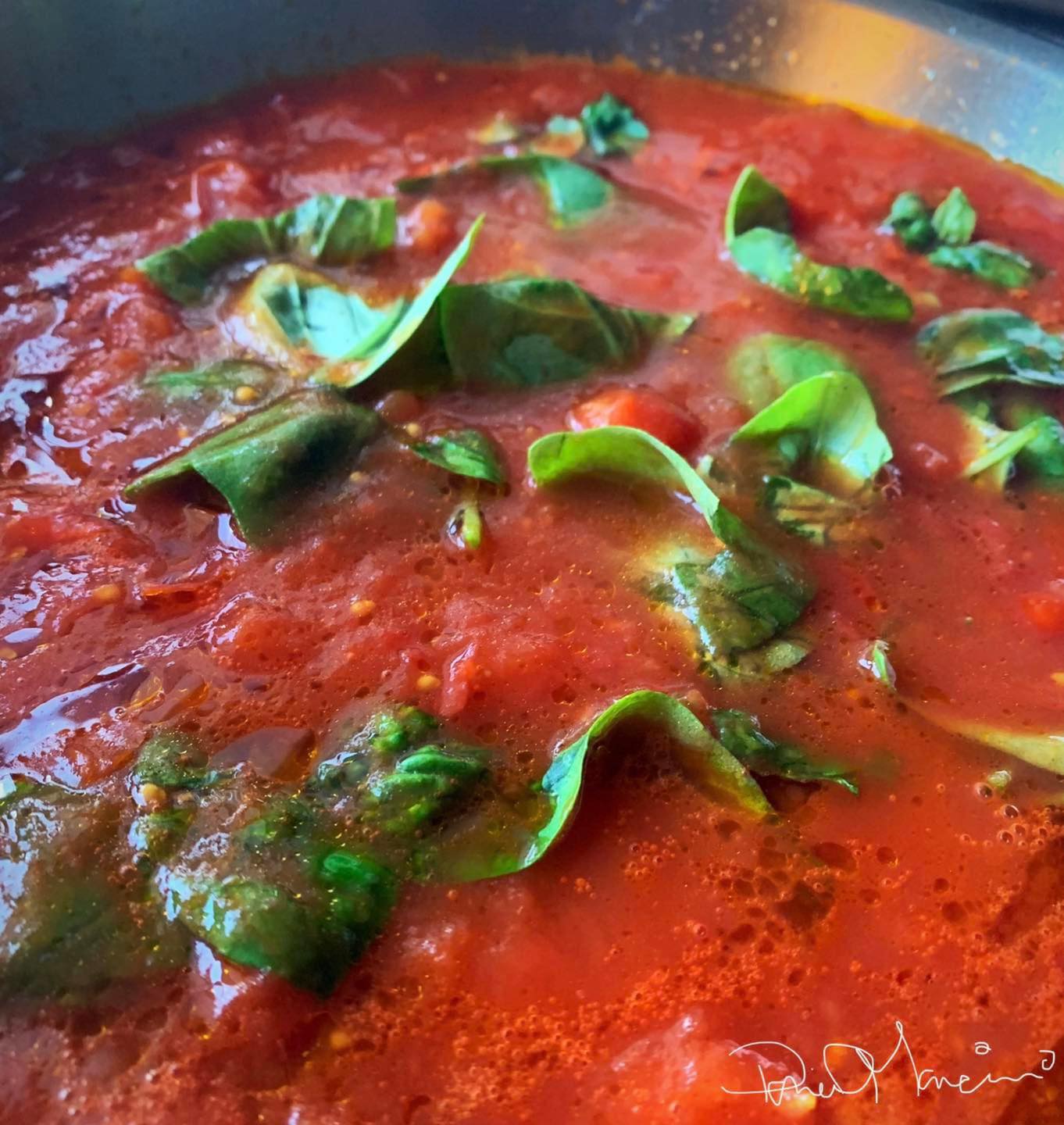 A close-up view of a skillet containing a rich red tomato sauce, garnished with fresh basil leaves. The sauce appears to be simmering, with visible hints of chopped tomatoes and seasoning.