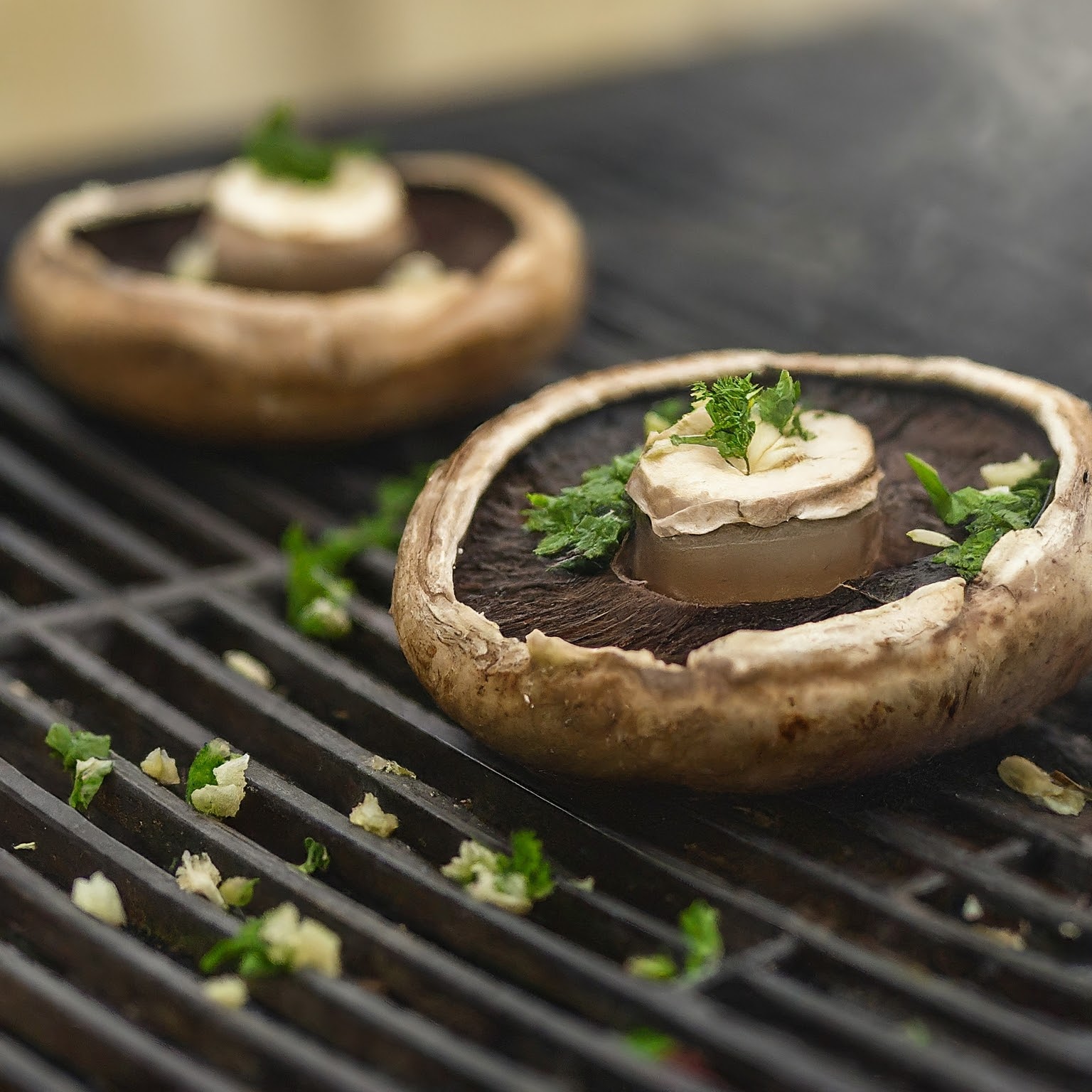 Close-up of two large portobello mushrooms on a grill, each topped with a knob of garlic herb butter and garnished with chopped fresh parsley. The grill grates and scattered bits of garlic and parsley can be seen around the mushrooms.