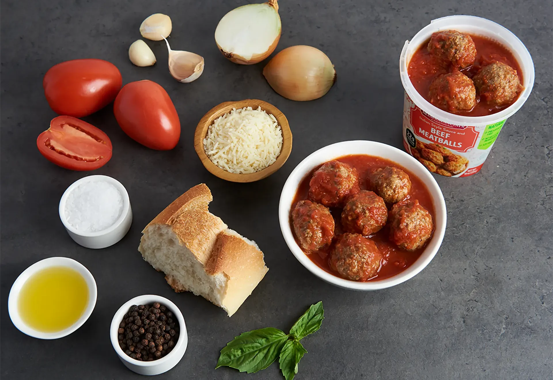 A bowl of beef meatballs in Italian style sauce sits on a dark surface surrounded by ingredients: whole and sliced tomatoes, onions, garlic cloves, grated cheese, a cup of olive oil, a bread piece, salt, pepper, basil leaves, and a container of the meatball product. _MamaMancinis