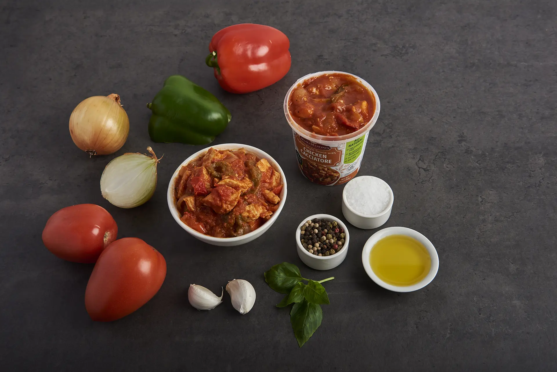 An assortment of ingredients for a chicken dish: two tomatoes, three bell peppers (red and green), two onions, garlic cloves, basil leaves, a bowl of seasoned meat or vegetable stew, peppercorns, salt, and olive oil on a dark surface. Perfect for creating an enticing Chicken Cacciatore. _MamaMancinis