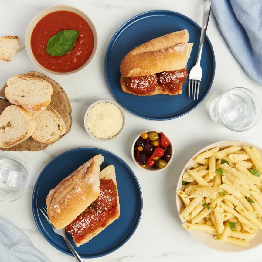 Two plates with beef meatball subs and a bowl of penne pasta topped with Italian style sauce and cheese are on a table. Accompanied by sliced bread, olives, peppers, and a bowl of tomato sauce, the spread is set on a light marble surface with a blue napkin and two glasses of water. _MamaMancinis