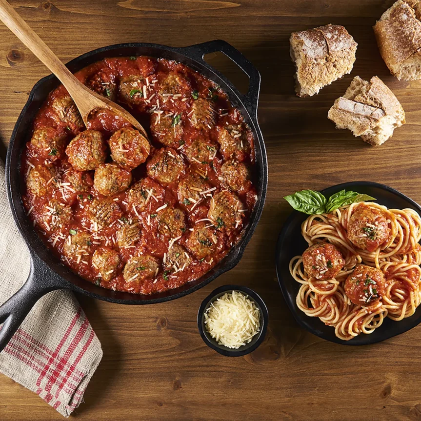 A cast iron skillet with beef meatballs in Italian style sauce, garnished with Parmesan. A plate of spaghetti with meatballs sits nearby, accompanied by grated cheese in a small bowl and slices of rustic bread on a wooden table. _MamaMancinis