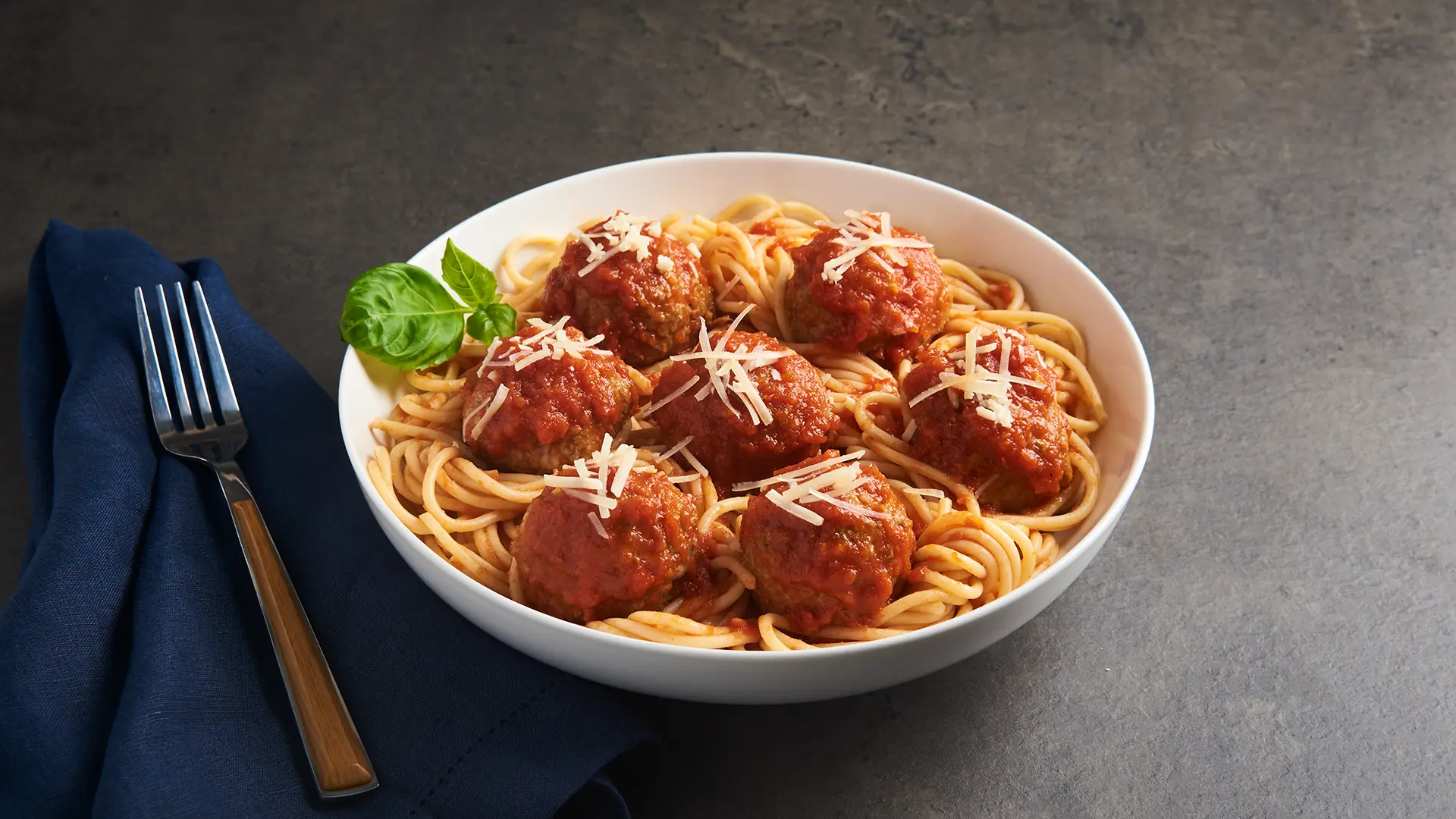 A bowl of spaghetti topped with beef meatballs in Italian-style sauce, garnished with grated cheese and a basil leaf. A fork rests on a dark blue napkin beside the bowl, set against a dark, textured surface. _MamaMancinis