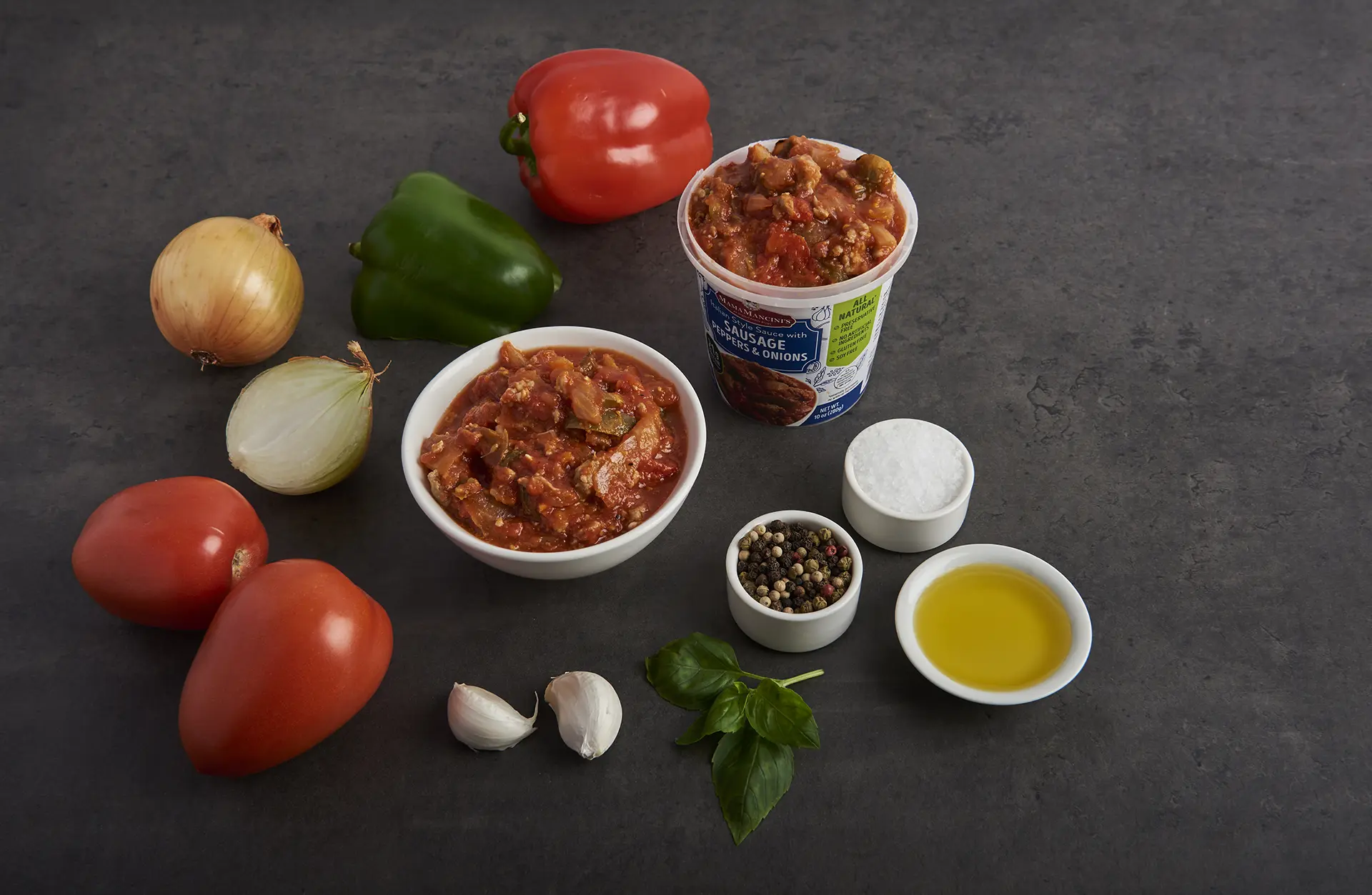 An arrangement of ingredients on a dark surface features diced tomatoes in a cup and a bowl, whole tomatoes, onions, garlic cloves, red and green peppers, basil leaves, small bowls of mixed peppercorns, salt, olive oil, and slices of savory sausage. _MamaMancinis