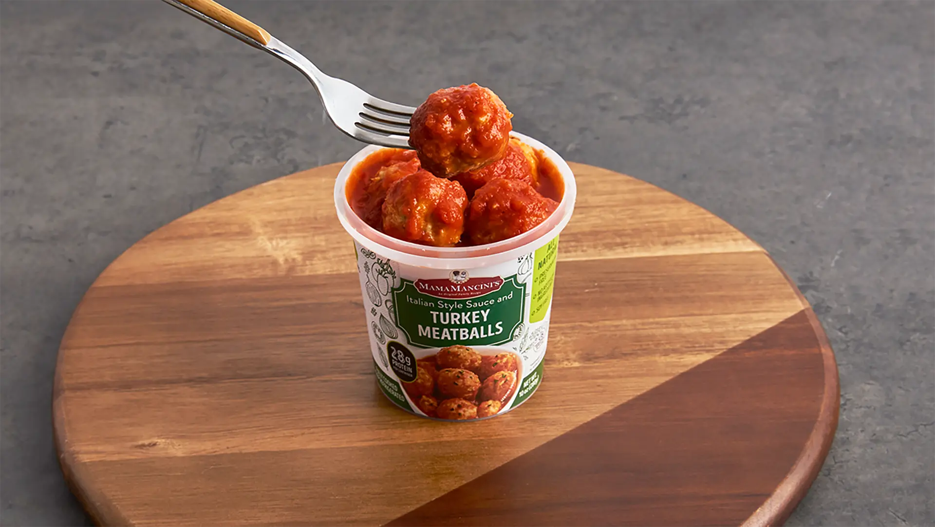 A fork lifts turkey meatballs above a cup labeled Italian Style Sauce with Turkey Meatballs. The cup rests on a round wooden board, set against a gray background. _MamaMancinis