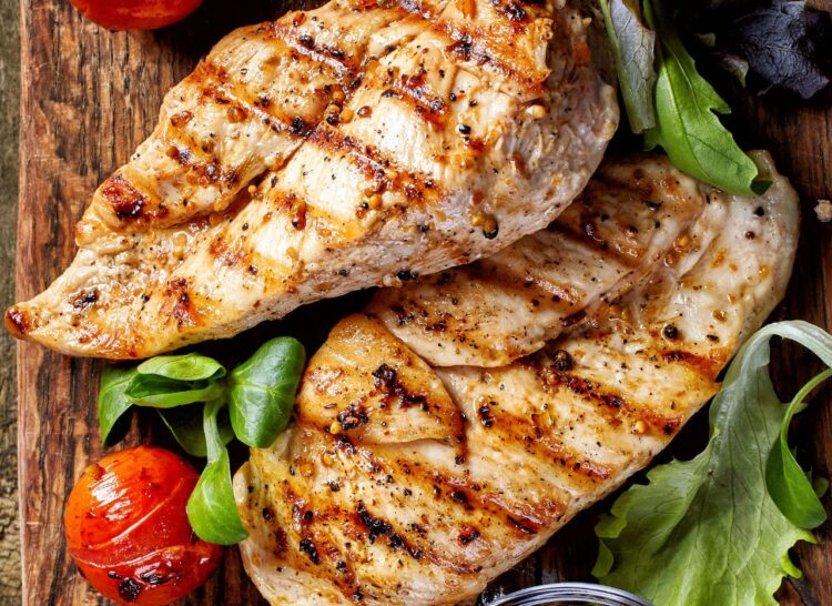 Grilled chicken breasts with char marks, garnished with leafy greens and roasted cherry tomatoes, served on a wooden cutting board. _MamaMancinis