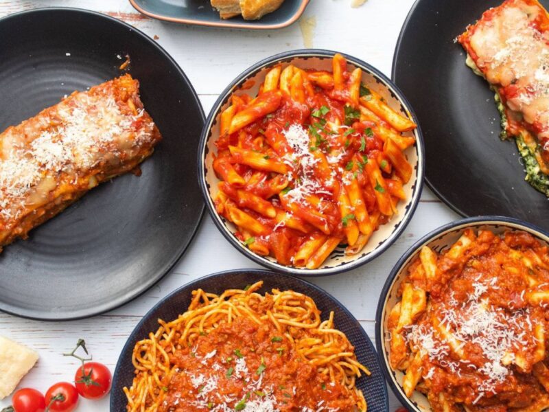 A variety of pasta dishes are on display, including spaghetti, penne, and lasagna, all topped with red sauce and cheese. The plates are arranged on a white table, with a cluster of cherry tomatoes nearby. _MamaMancinis