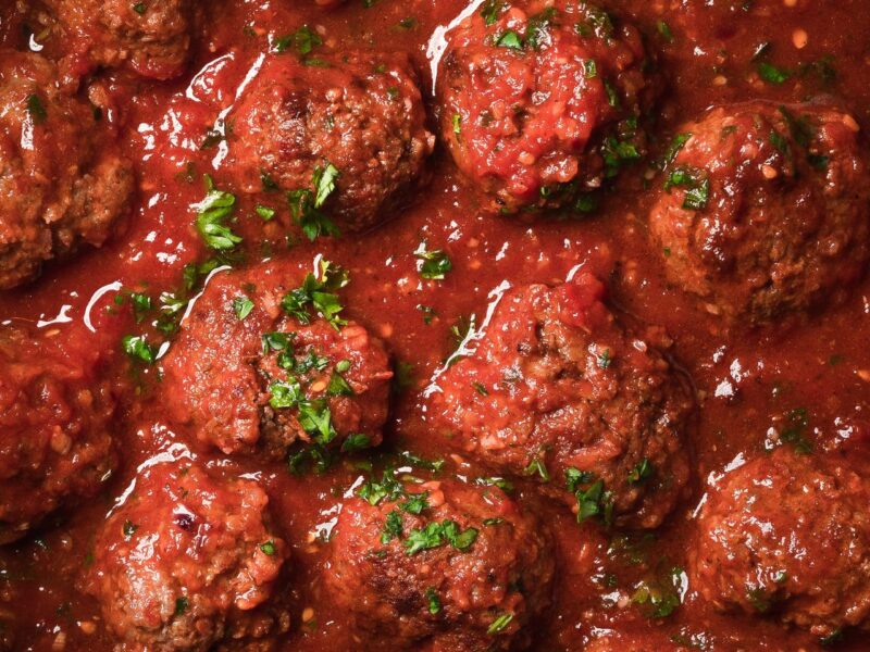 Close-up of meatballs garnished with chopped parsley, simmering in a rich tomato sauce. _MamaMancinis