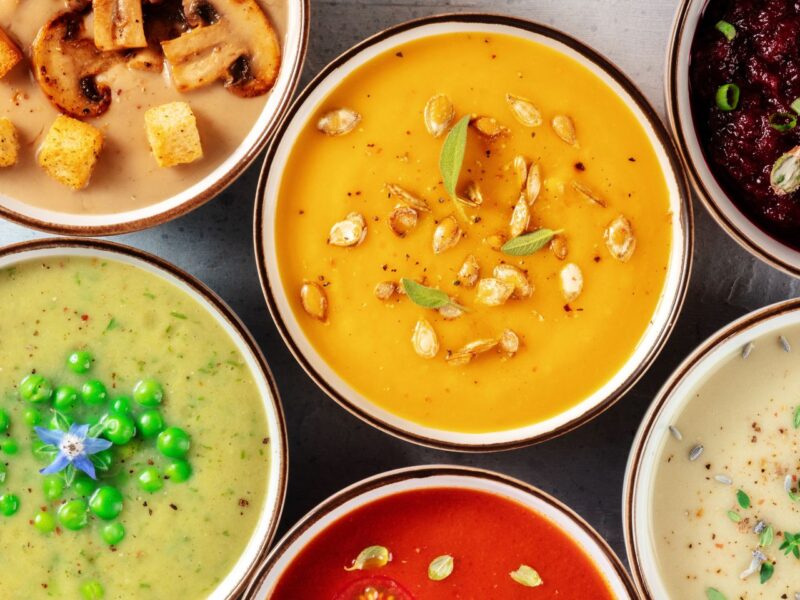 A variety of colorful soups in bowls are arranged in a circle. Each soup is garnished uniquely with items like seeds, herbs, peas, and croutons, showcasing different textures and ingredients. _MamaMancinis