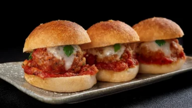 Three meatball sliders sit perfectly on a rectangular plate, showcasing meatballs with melted cheese and tomato sauce. Topped with small parsley leaves, they're nestled in round golden buns, all set against a dark background. _MamaMancinis