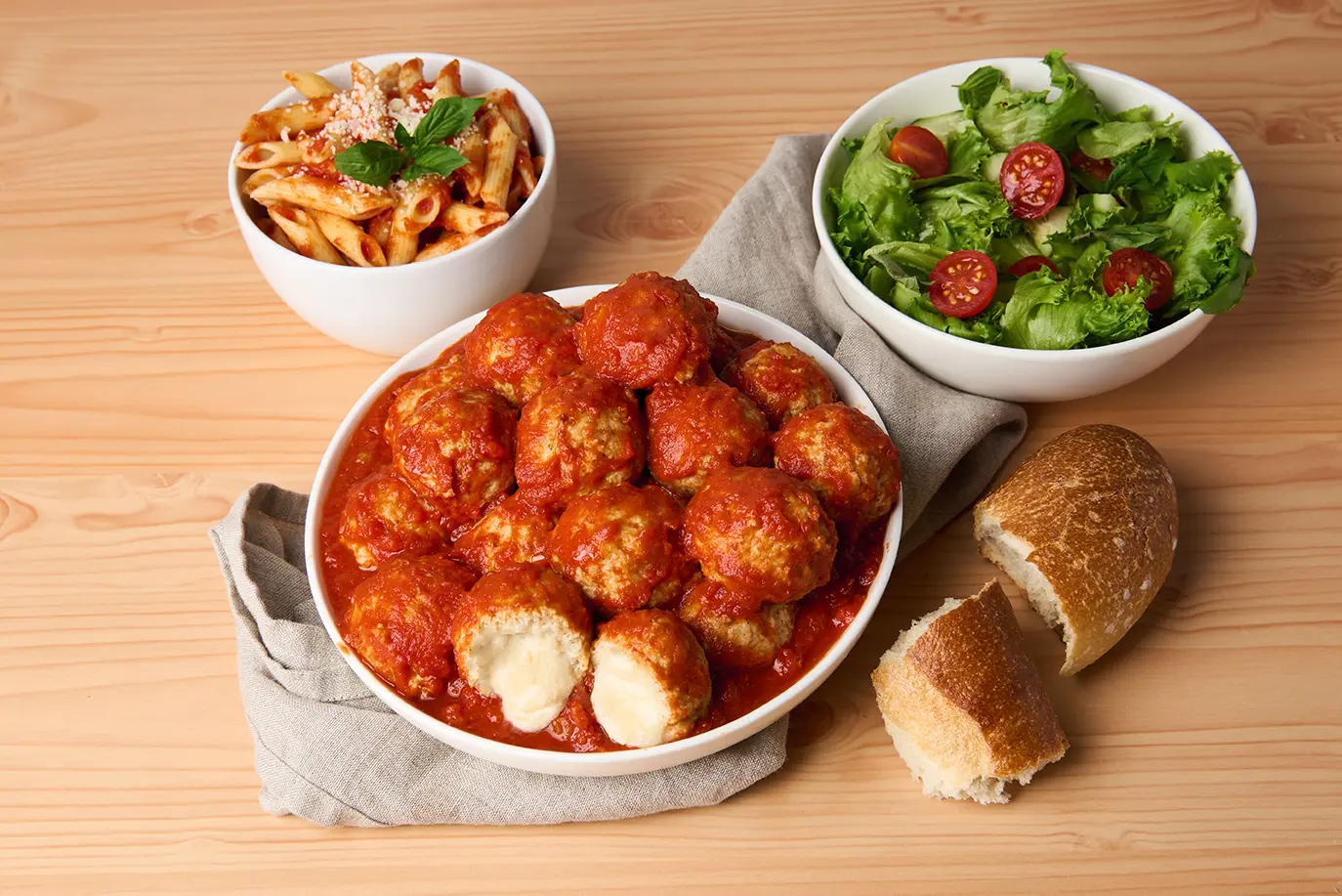 A plate of Cheese Stuffed Chicken Meatballs in Italian Style Sauce takes center stage, paired with a piece of crusty bread. In the background, a bowl of penne pasta with cheese and herbs sits to the left, while a salad with cherry tomatoes and lettuce adorns the right. _MamaMancinis