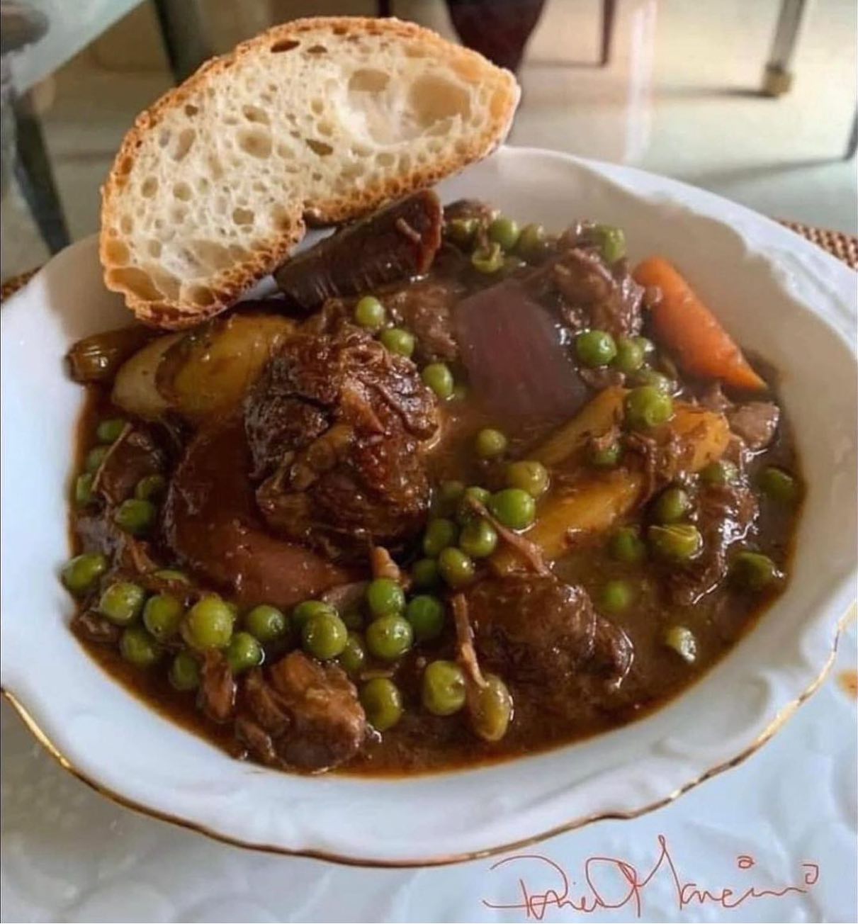 A bowl of beef stew filled with chunks of beef, peas, potatoes, carrots, and onions in a rich gravy with a hint of garlic. A slice of crusty bread is resting on the edge of the bowl. _MamaMancinis