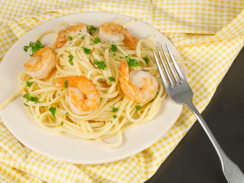 A plate of linguini topped with shrimp and garnished with parsley is on a table. There's a silver fork placed beside the dish. The background features a yellow and white checkered fabric, adding a touch of charm to this delightful shrimp scampi setting. _MamaMancinis