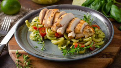 A ceramic plate showcases flame-grilled chicken breast slices atop a bed of pasta with pesto sauce, cherry tomatoes, and fresh herb garnish. A fork and lime rest nearby on the rustic wooden table. _MamaMancinis