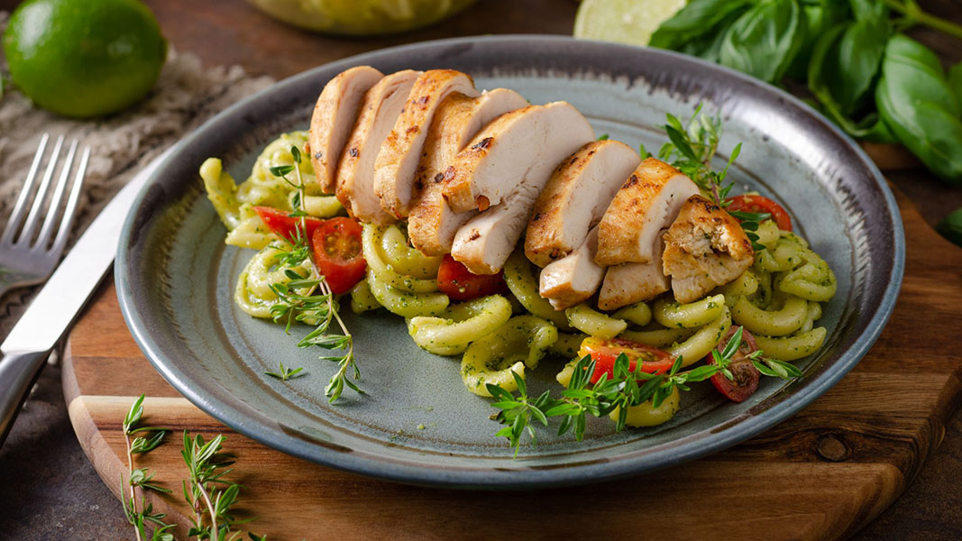 A ceramic plate showcases flame-grilled chicken breast slices atop a bed of pasta with pesto sauce, cherry tomatoes, and fresh herb garnish. A fork and lime rest nearby on the rustic wooden table. _MamaMancinis