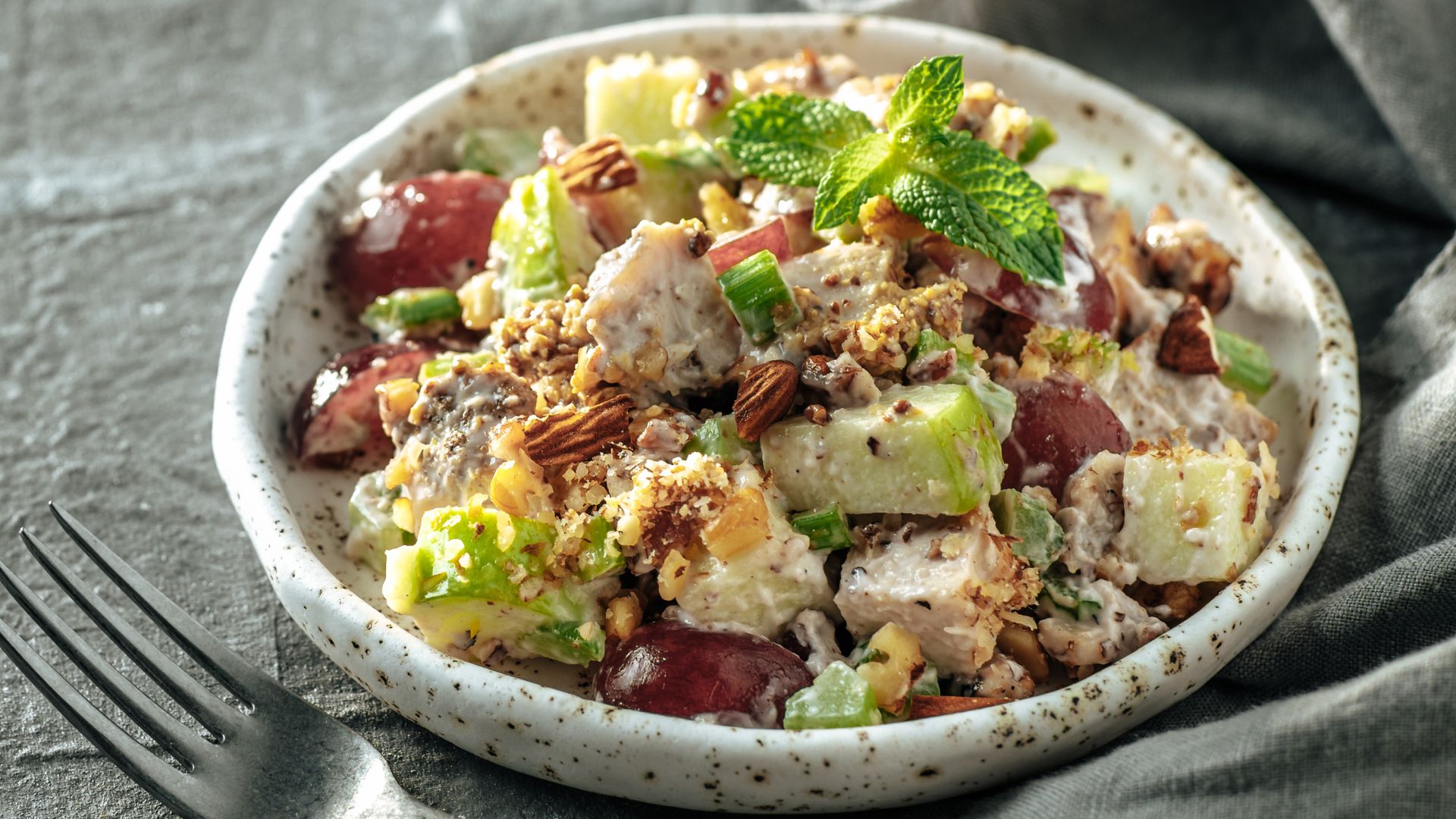 A plate of Waldorf salad featuring diced apples, grapes, celery, and walnuts, garnished with fresh mint leaves. This creamy salad is complemented by flame-grilled chicken pieces and served on a speckled plate, with a fork nearby on a textured gray cloth. _MamaMancinis