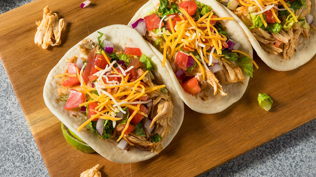 Three flame-grilled tacos rest on a wooden board, brimming with tender shredded chicken, crisp lettuce, diced tomatoes, and red onions. Each taco is generously topped with shredded cheese and aligned side by side on a speckled countertop. _MamaMancinis