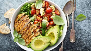 A fresh Chicken and Avocado Salad bowl features flame grilled chicken slices, halved avocados, cherry tomatoes, and mozzarella balls. Basil leaves are sprinkled on top, served with bread slices on the side. A fork and spoon rest nearby. _MamaMancinis