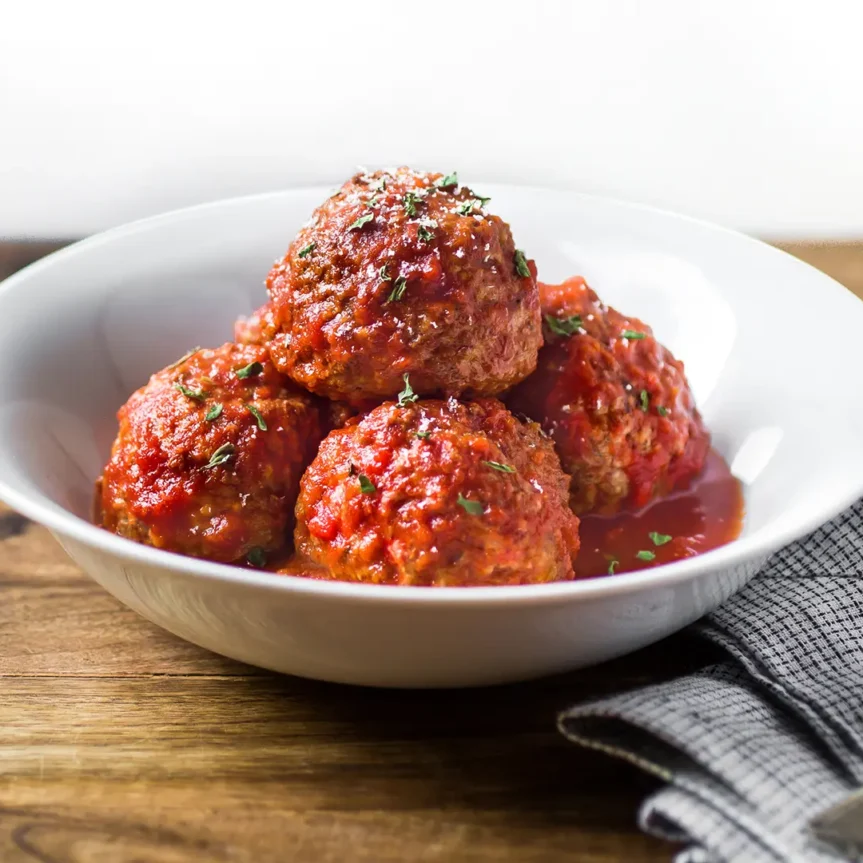 A white bowl filled with four jumbo beef meatballs covered in Italian style sauce, garnished with herbs. The bowl is set on a wooden table next to a gray checked napkin and a fork—just like you'd find at Costco. _MamaMancinis