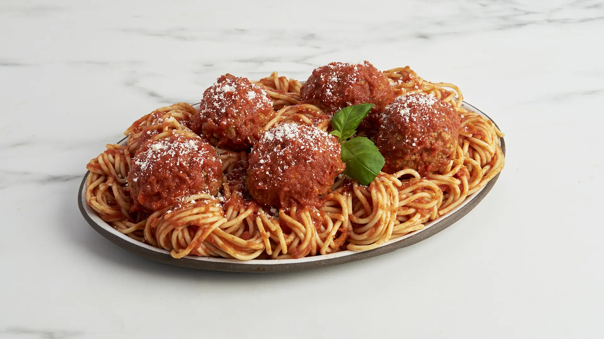 A plate of spaghetti topped with jumbo beef meatballs, smothered in Italian style sauce and garnished with grated cheese. A sprig of fresh basil sits in the center. This delectable dish is set on a pristine white marble surface. _MamaMancinis