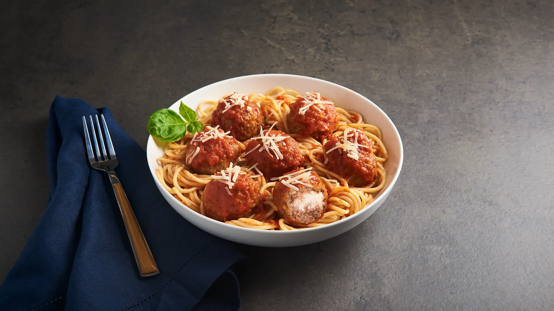 A bowl of spaghetti topped with five cheese-stuffed meatballs and grated cheese, garnished with a basil leaf. A fork rests on a dark blue napkin next to the bowl, set against a dark, textured background. _MamaMancinis