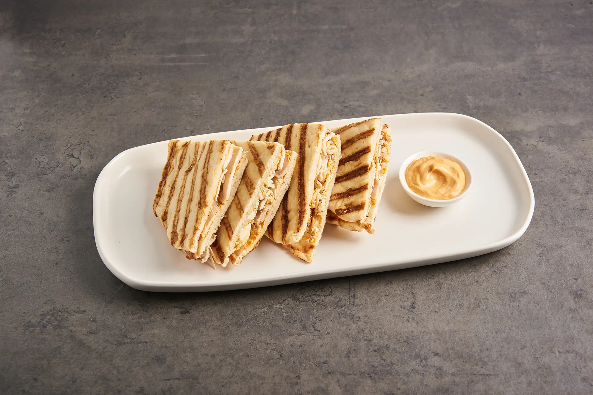 Grilled quesadilla slices, sprinkled with a hint of Parmesan, are neatly arranged on an oval white plate, accompanied by a small cup of orange dipping sauce. The table surface is a dark gray stone. _MamaMancinis