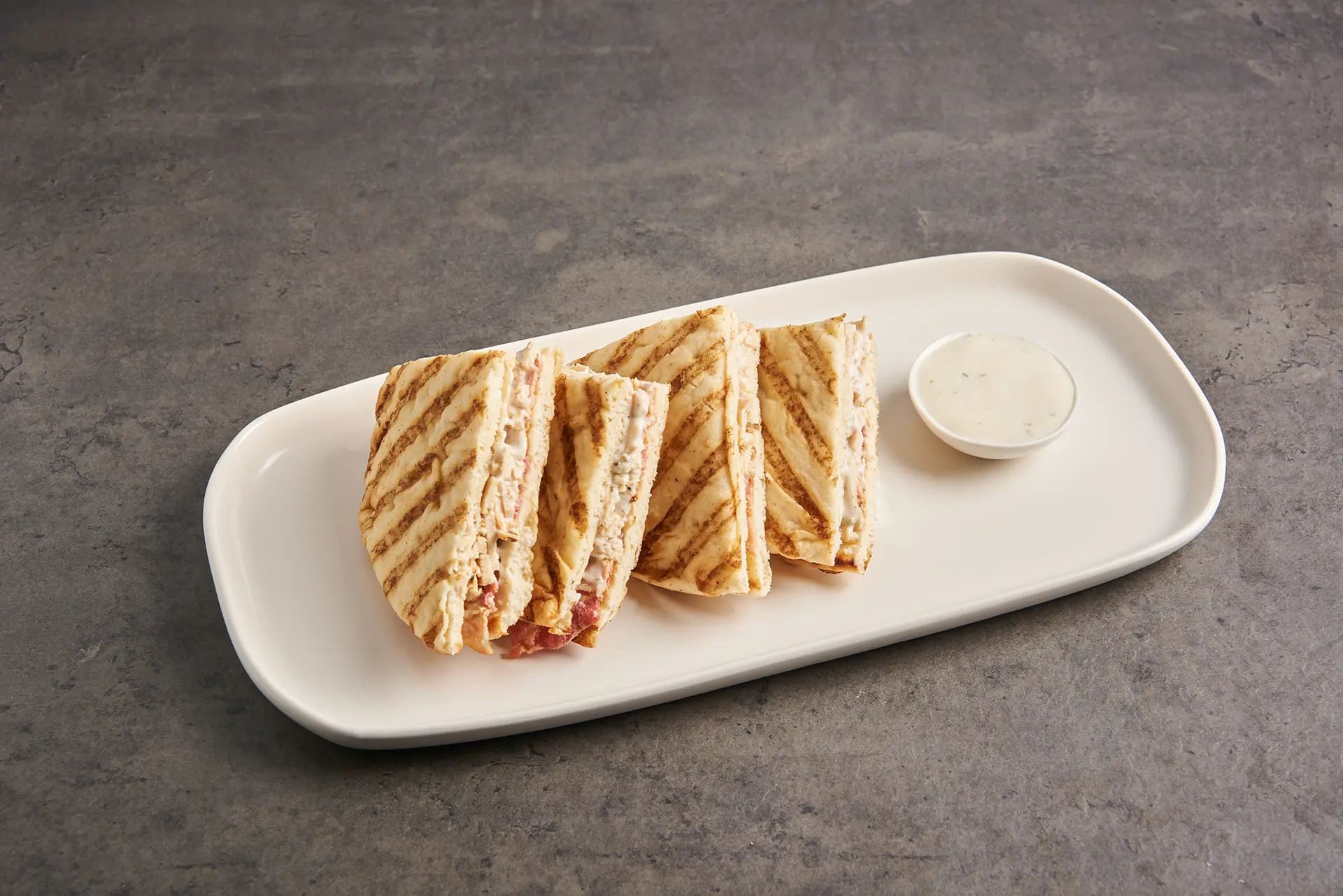 A rectangular white plate holds four triangular slices of grilled Parmesan sandwich with filling, arranged neatly. A small cup of creamy dipping sauce is placed beside the sandwiches. The background is a gray textured surface. _MamaMancinis