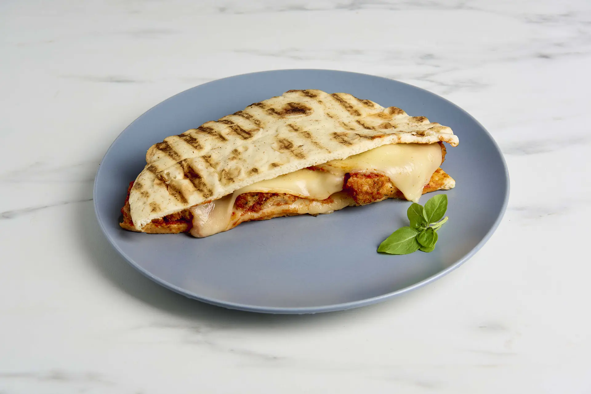 A grilled piadina folded in half, filled with melted cheese and tender grilled chicken, is served on a blue plate. A sprinkle of Parmesan adds flavor, while a sprig of fresh basil rests beside it on the white marble surface. _MamaMancinis