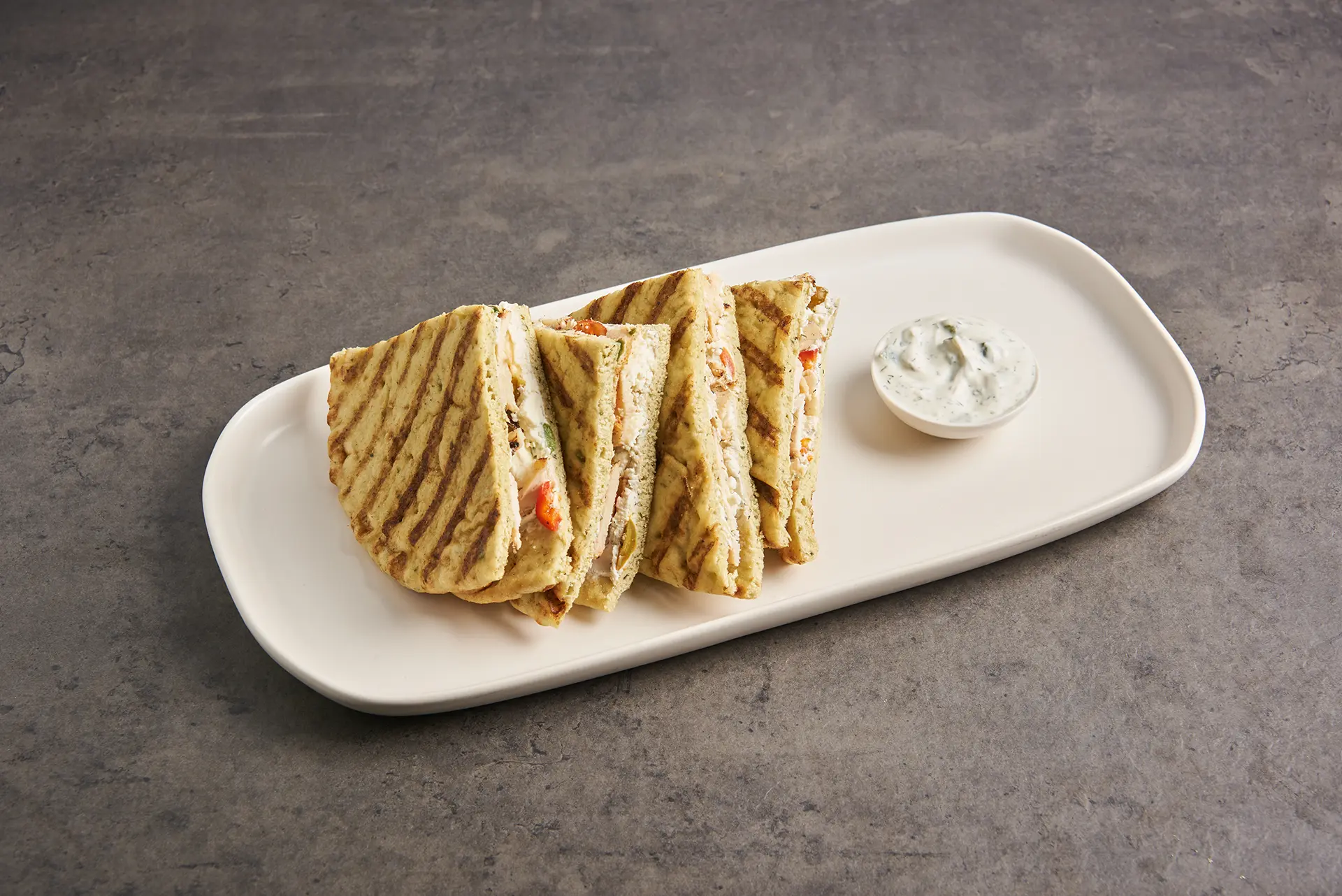 A grilled quesadilla cut into triangles is served on an oval white plate, accompanied by a small dish of creamy dipping sauce. Parmesan is sprinkled throughout the vegetable and cheese filling, adding a savory twist as it rests on a gray, textured surface. _MamaMancinis