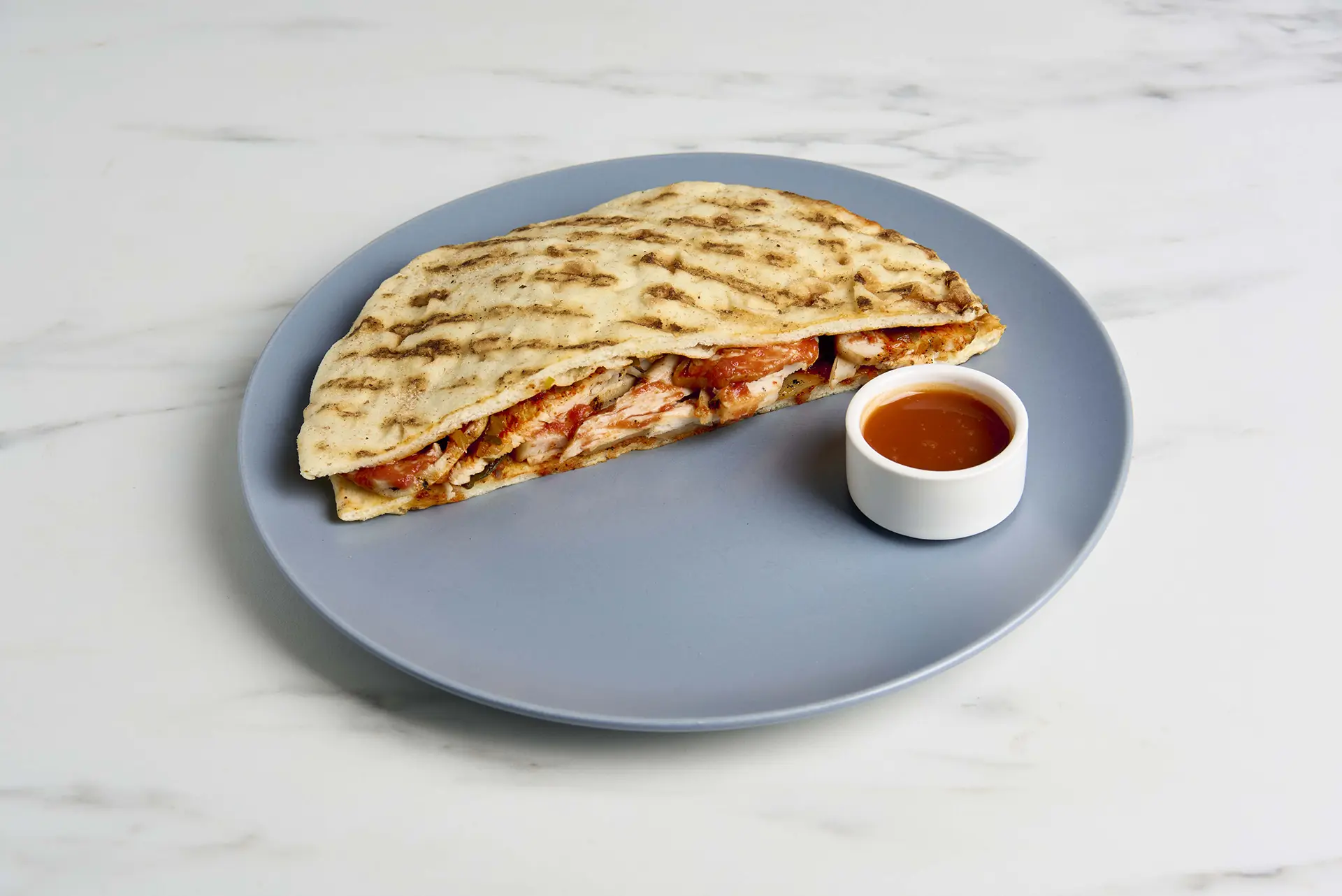 A grilled chicken pita sandwich filled with sliced chicken, vegetables, and a sprinkle of Parmesan on a grey plate, accompanied by a small white cup of dipping sauce on a marble surface. _MamaMancinis