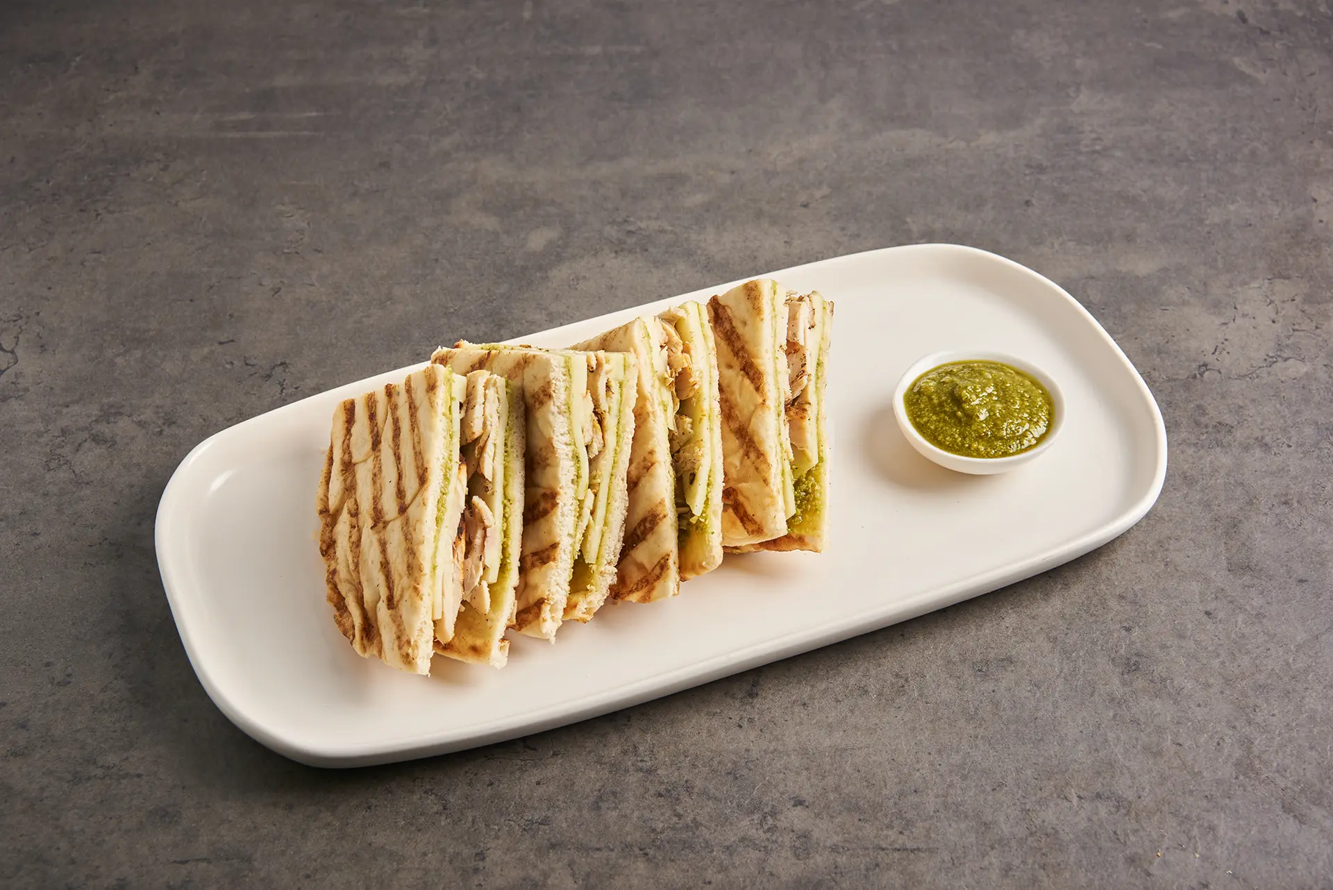A white rectangular platter displays grilled tortilla wedges filled with melted cheese, minced herbs, and a hint of Parmesan. A small dish of green sauce accompanies the savory quesadillas, all set against a textured gray surface. _MamaMancinis