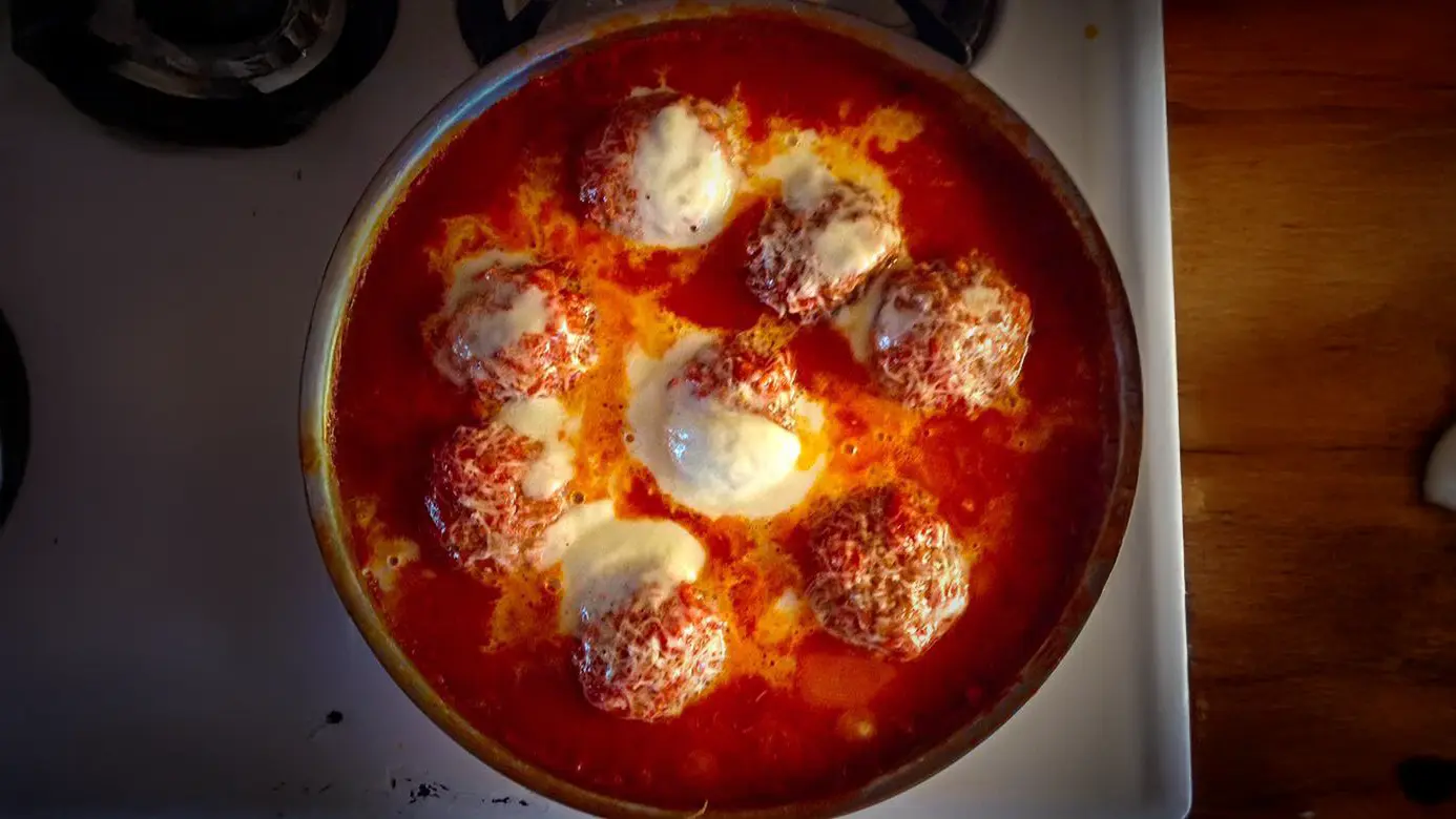 A skillet with meatballs simmering in a rich tomato sauce, topped with melted mozzarella cheese, sits on the stovetop. The golden-brown meatball parmigiana is partially submerged in the vibrant, bubbling sauce, creating an appetizing display. _MamaMancinis