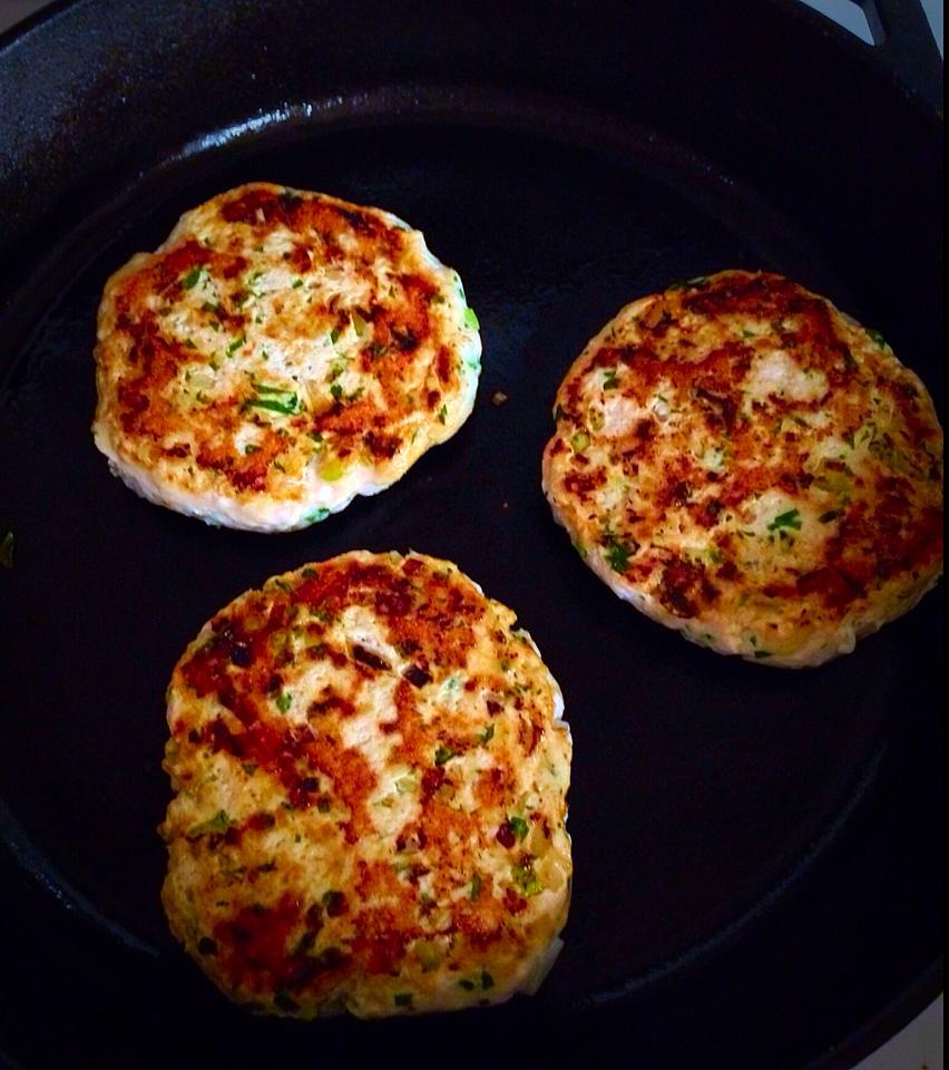 Three golden-brown turkey burgers sizzling in a black cast iron skillet. The patties are speckled with green herbs, indicating they may be seasoned. The image captures the appetizing texture and color of the patties while they cook. _MamaMancinis