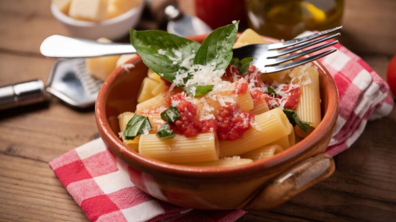 A rustic bowl of rigatoni pasta is adorned with filetto di pomodoro, fresh basil, and grated Parmesan cheese on a wooden table. A fork rests atop the bowl, alongside a red and white checkered cloth. In the background, Parmesan and oil complete this rustic style setting. _MamaMancinis