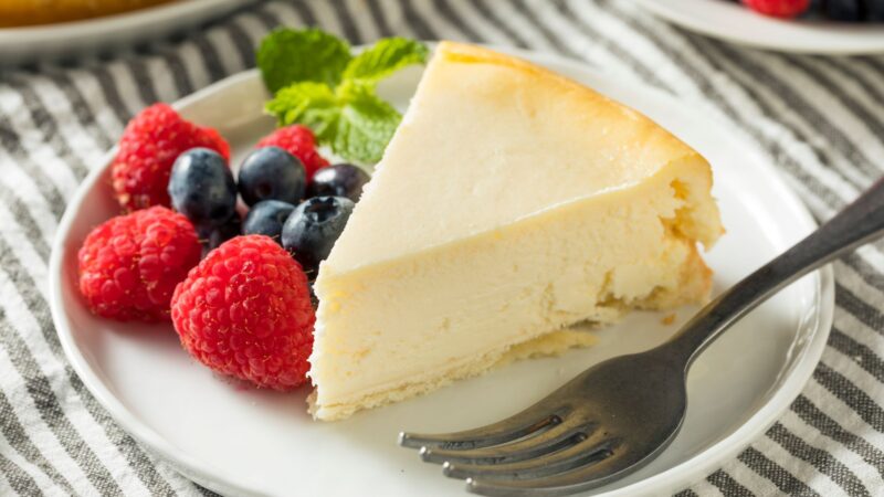 A slice of cheesecake made with Italian ricotta cheese sits on a white plate, garnished with fresh raspberries, blueberries, and a sprig of mint. A fork lies beside the cake on a striped cloth background. _MamaMancinis