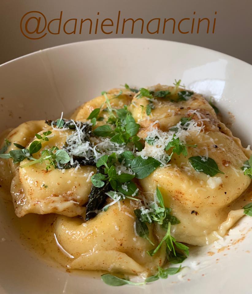 A bowl of ravioli garnished with fresh herbs and grated cheese, served in a luscious sage brown butter sauce. The dish is presented in a white bowl, and the text @danielmancini is visible above. _MamaMancinis