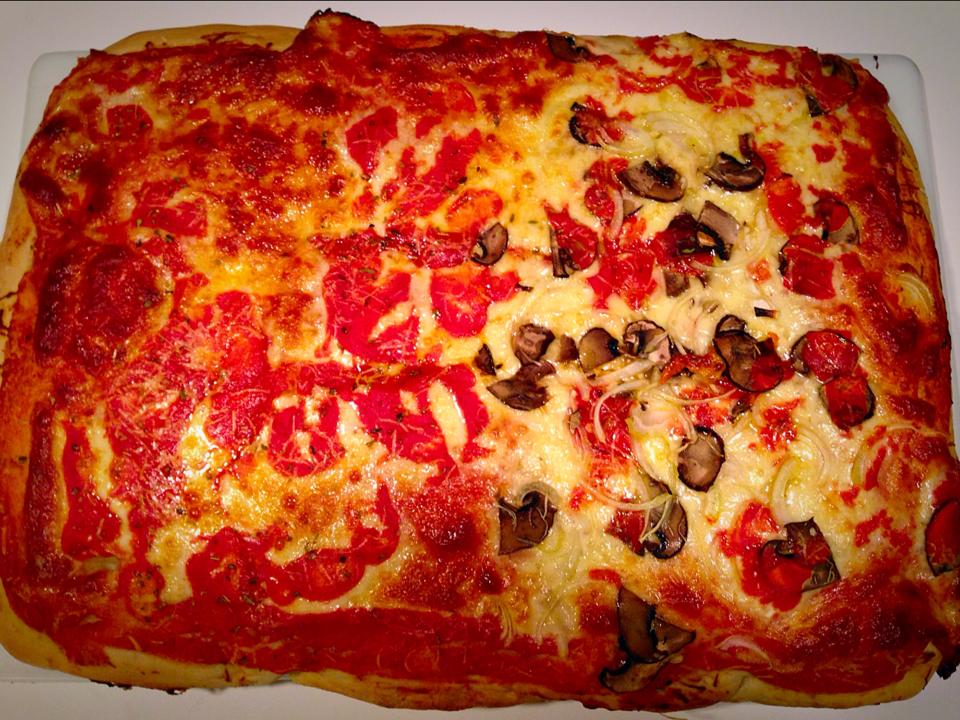 A rectangular Sicilian homemade pizza with a golden-brown crust, topped with melted cheese, tomato sauce, slices of mushroom, and bits of onion. The toppings are unevenly distributed across the surface. _MamaMancinis