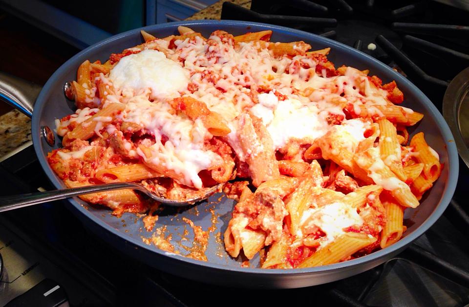 A close-up of a skillet showcases Stove Top Baked Ziti, where penne pasta mingles with rich tomato sauce and savory ground meat, all crowned with melted cheese and ricotta dollops. A spoon rests invitingly inside the skillet on the stovetop, ready to serve this comforting dish. _MamaMancinis