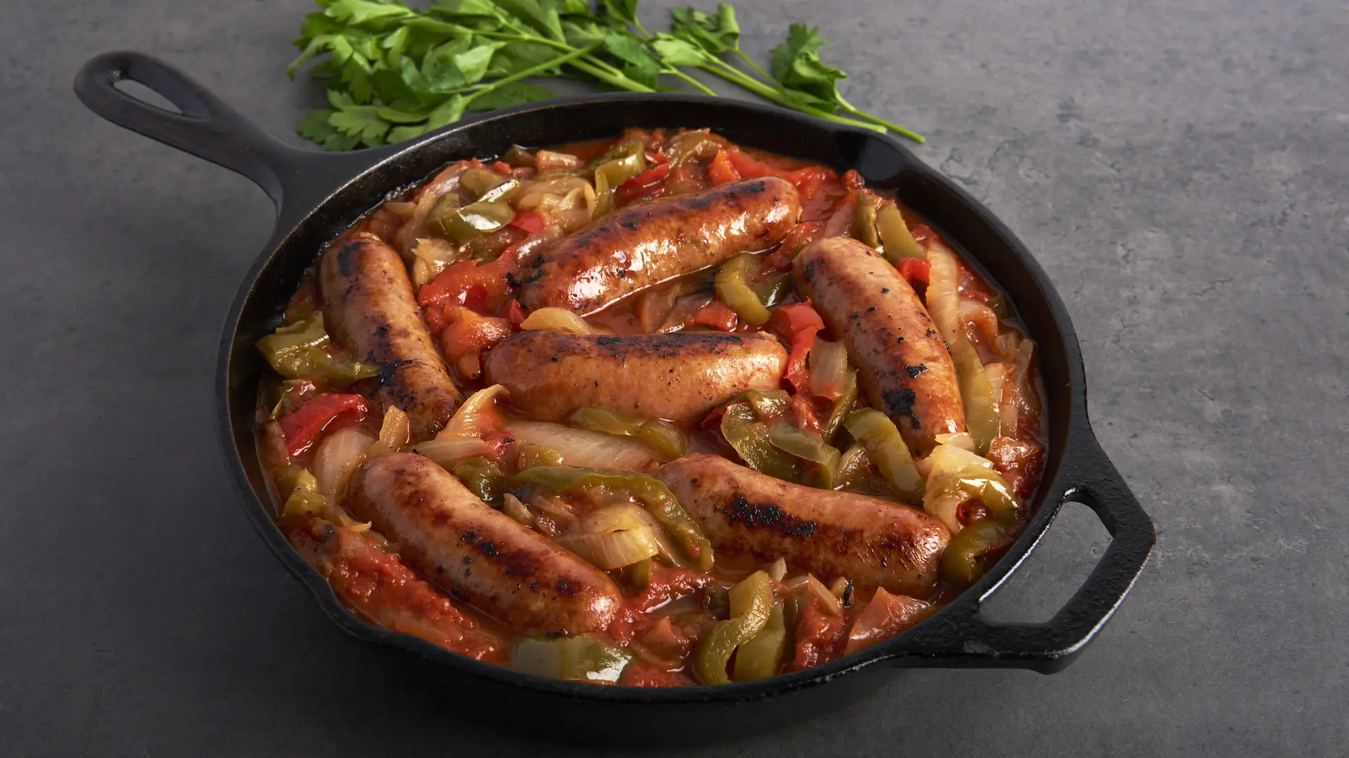 A cast iron skillet with browned Costco sausages, sliced green and red peppers, and onions in a savory Italian style sauce. Fresh parsley is placed beside the skillet, all set on a dark gray surface. _MamaMancinis