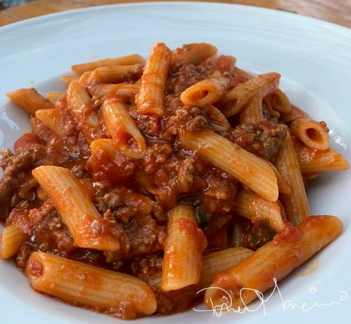 A white plate filled with penne pasta topped with a rich tomato and ground meat sauce, garnished with herbs and tender eggplant. The dish appears freshly served and appetizing. _MamaMancinis