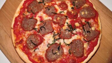 A cooked meatball pizza topped with tomato sauce, cheese, and herbs sits on a wooden pizza peel against a plain white background. _MamaMancinis