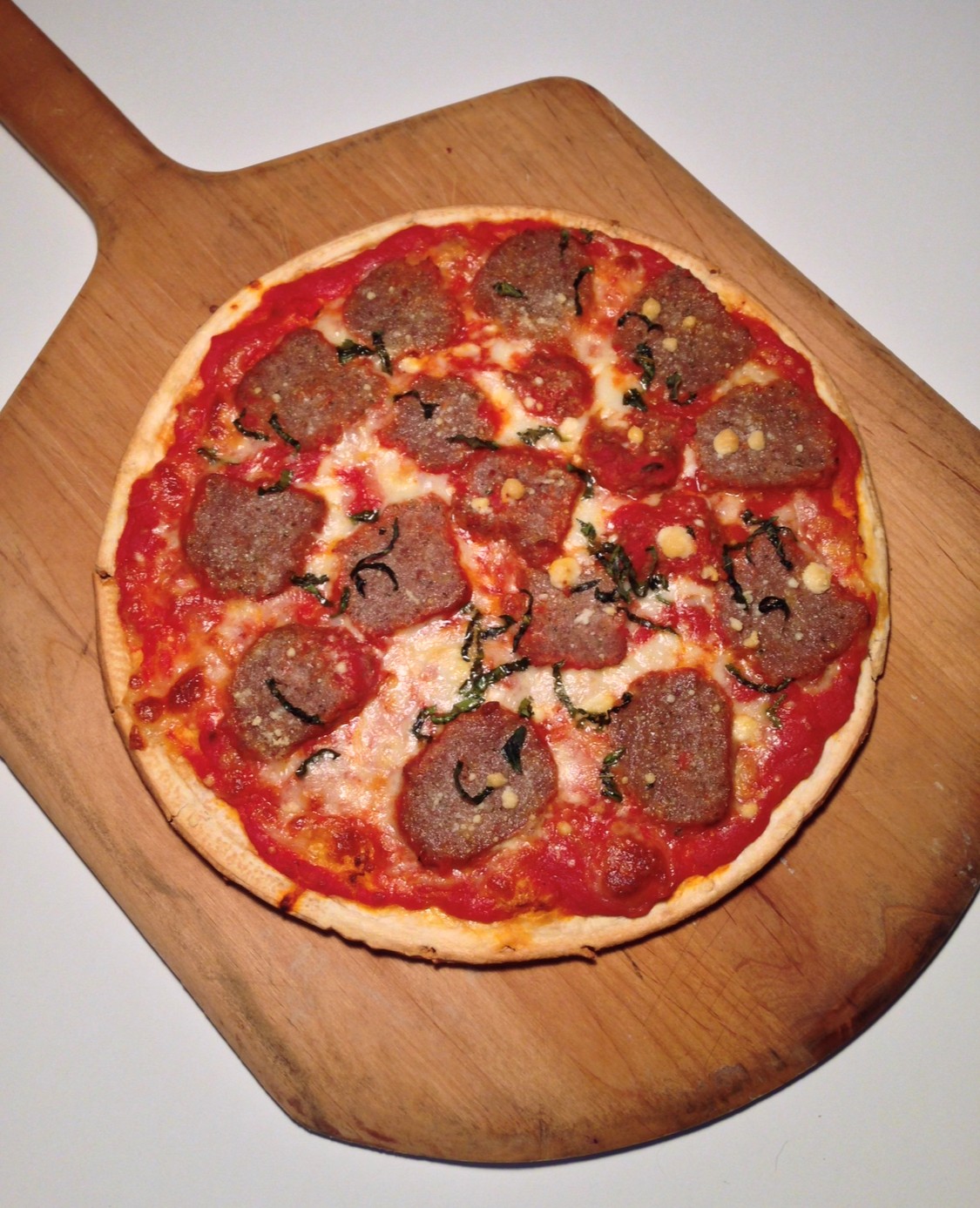 A cooked meatball pizza topped with tomato sauce, cheese, and herbs sits on a wooden pizza peel against a plain white background. _MamaMancinis