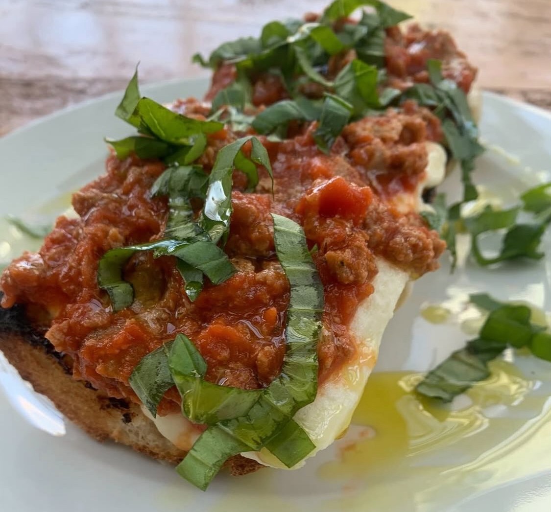 A close-up of toasted bread topped with melted cheese, meat and tomato sauce, fresh basil strips, and a drizzle of olive oil on a white plate—perfect alongside a warm bowl of soup. _MamaMancinis